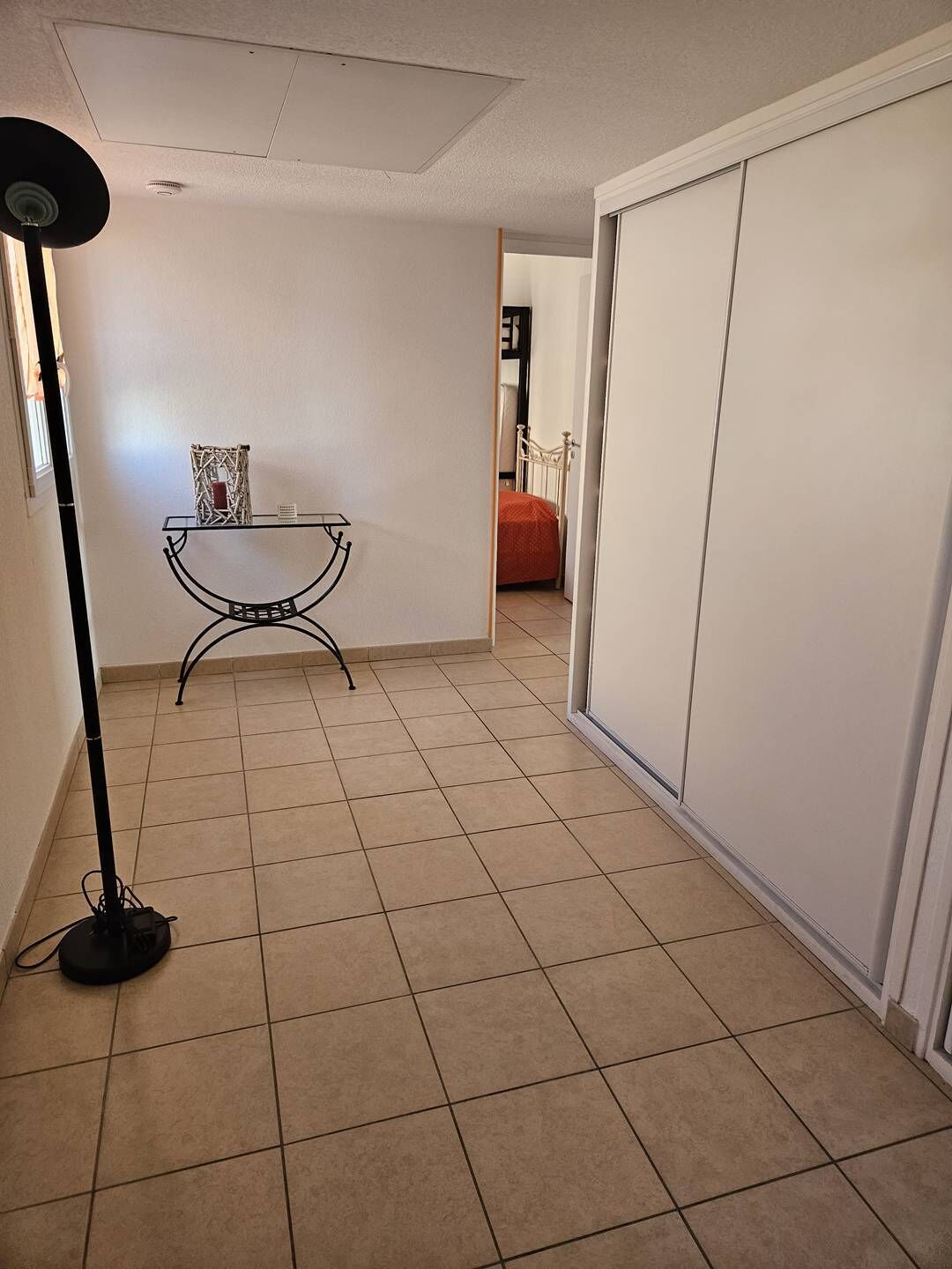 Un couloir avec un sol carrelé, un lampadaire, une petite table ornée d’un objet décoratif et, au fond, un lit recouvert d’une couverture orange.
