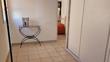 Un couloir avec un sol carrelé, un lampadaire, une petite table ornée d’un objet décoratif et, au fond, un lit recouvert d’une couverture orange.