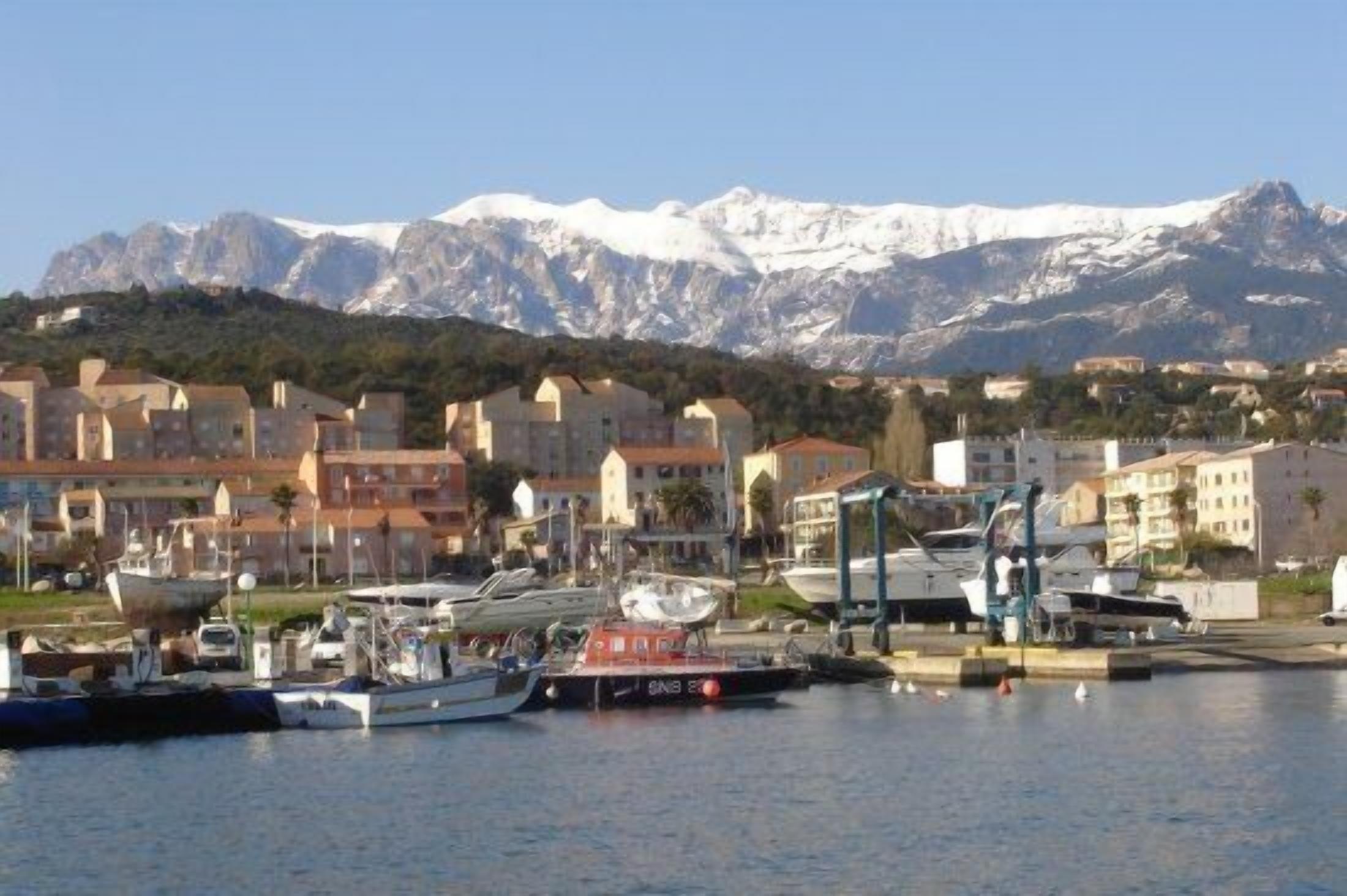 A marina with boats docked, a mountainous backdrop, and buildings with a Mediterranean architectural style.
