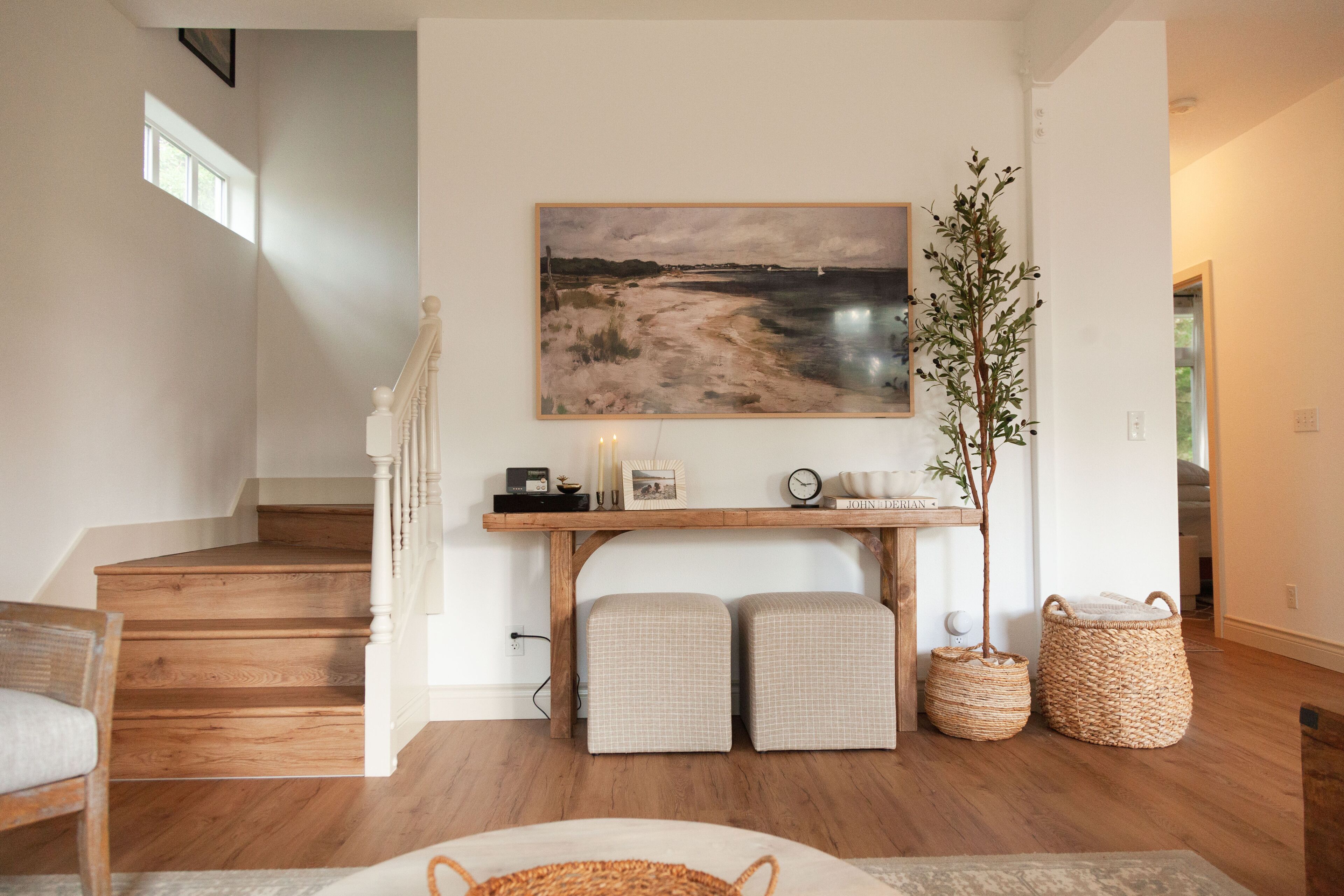 Un escalier avec des marches en bois et une rampe blanche, une console sur laquelle est posée une peinture, deux bancs rembourrés, des paniers en osier et une plante.