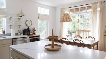 A kitchen with a marble countertop, wooden dining table, and chairs. There is a coffee maker, a clock, and a window with curtains.