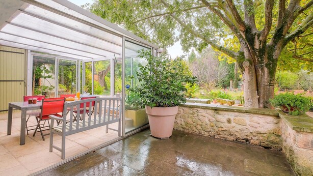 A covered patio with a table and chairs, a large potted plant, and a stone wall.