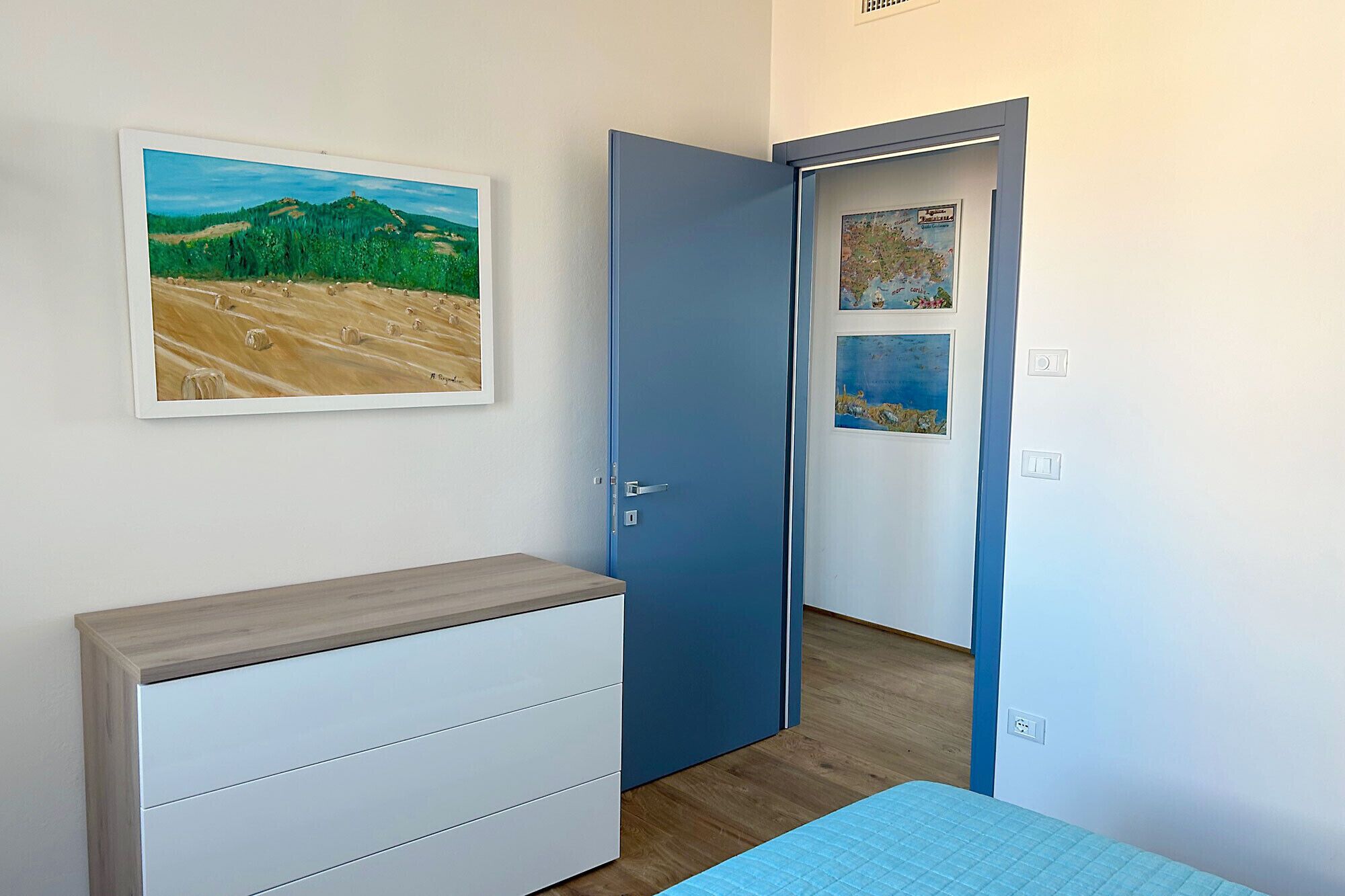 A bedroom with a wooden dresser, a bed with a blue quilt, a blue door, and a wall with a landscape painting.