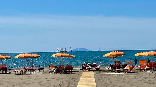 Ein Strand mit Liegestühlen und Sonnenschirmen, einem Sandweg und Segelbooten in der Ferne.