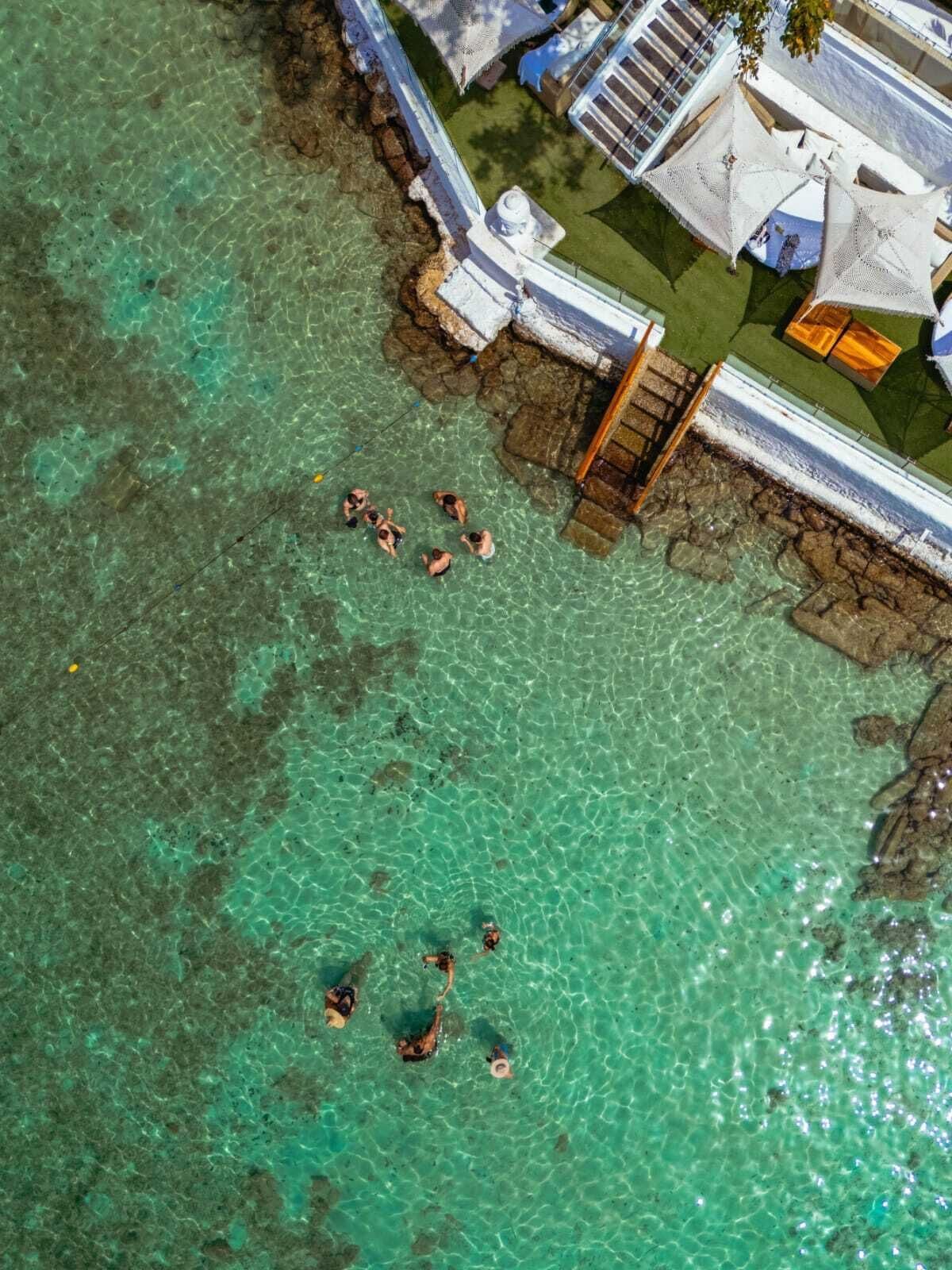 An aerial view of a coastal area with clear turquoise waters, a rocky shoreline, and a few people swimming. There are structures with white walls and a covered area with a roof.