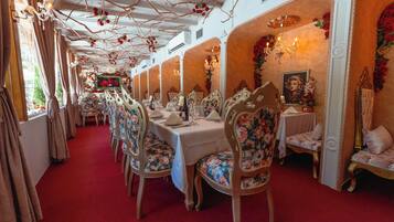 A long dining room with multiple tables and chairs, a red carpet, and decorative elements like chandeliers and floral arrangements.