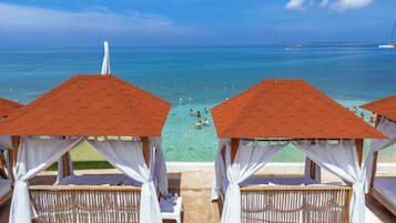 Blick auf einen Strand mit klarem Wasser, sandigem Ufer und Holzcabanas mit weißen Vorhängen.