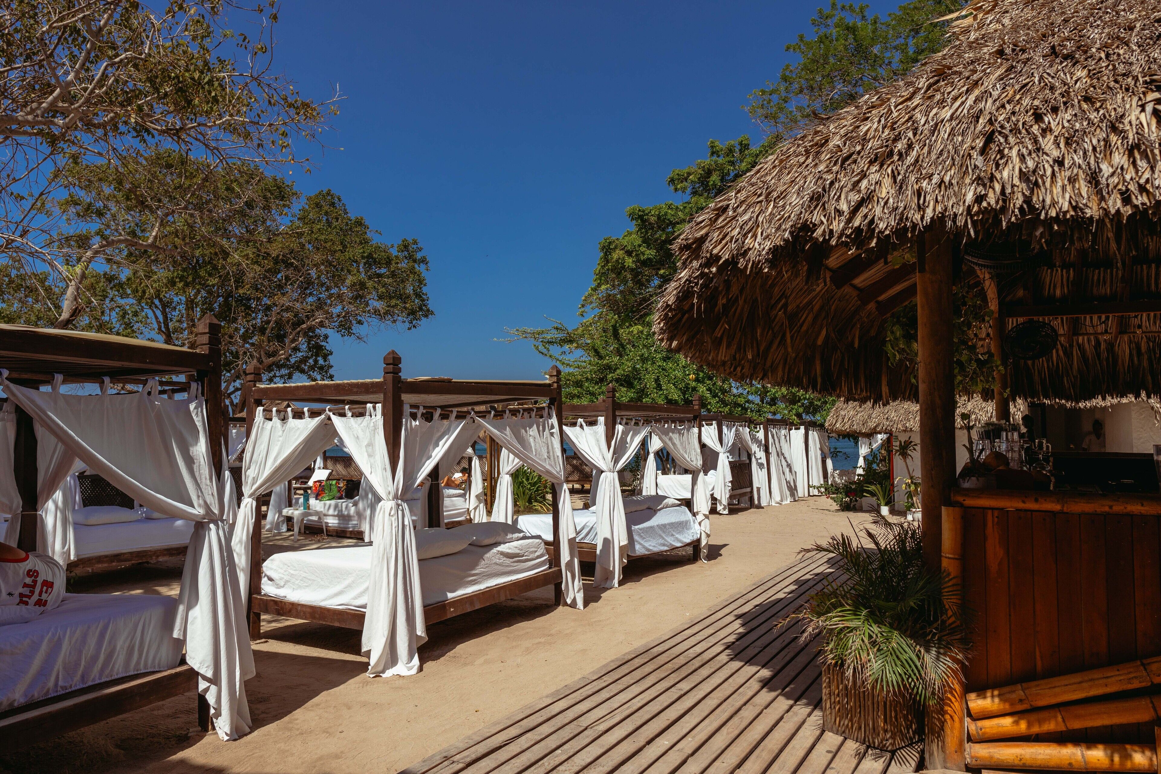 A beachside resort with thatched-roof huts and wooden beds with white canopies.