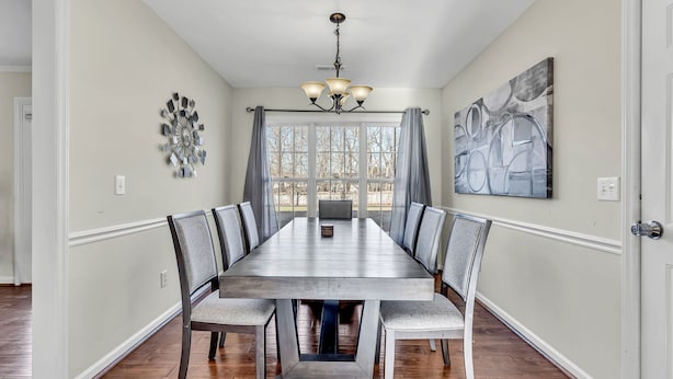 A dining room with a long table, chairs, a chandelier, a window with curtains, and a wall art piece.