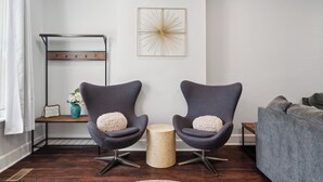 A modern living room with two dark armchairs, a grey sofa, a decorative wall piece, and a small wooden side table with a vase.