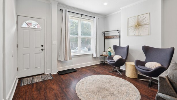 A modern living room with a white door, a large window with curtains, two dark armchairs, a round rug, and a decorative wall piece.