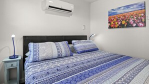 A bedroom with a large bed, a nightstand, an air conditioner, and a wall-mounted painting.