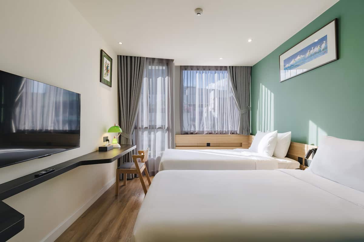 A hotel room with a large flat-screen TV, a desk with a lamp, two beds, a window with curtains, and a painting on the wall.