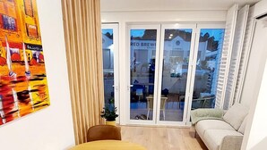 Una sala de estar moderna con una mesa redonda de madera, dos sillas marrones, un sofá beige y un gran ventanal con vista a un edificio.