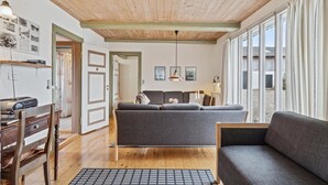 Una sala de estar acogedora con techo de madera, un sofá gris, una silla de madera y una mesa de centro. En la pared hay cuadros enmarcados y una ventana con cortinas.