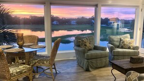 A room with a glass table and chairs, a view of a lake, and a sunset. - Cottage Time - Marsh Front in Garden City Beach, SC (Garden City)