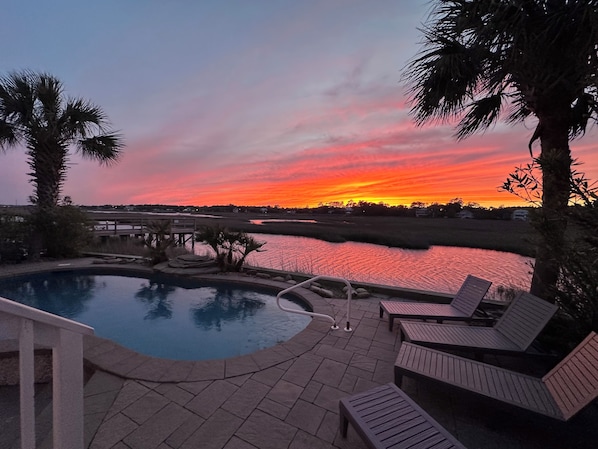 A swimming pool with lounge chairs, a wooden deck, and a view of a sunset over water.