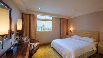 A hotel room with a bed, bedside table, lamp, chair, and a view of the city through the window.