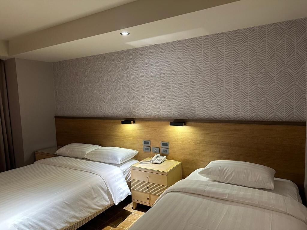 A hotel room with two beds, a wooden headboard, a patterned wall, and a nightstand with a phone.