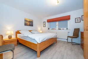 Ein Schlafzimmer mit einem hölzernen Bett, einem Sessel, einem Schreibtisch und einem Fenster mit Jalousien.