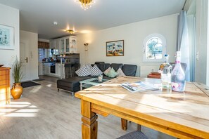 Ein modernes Wohnzimmer mit einem Holzesstisch, einer Couch mit Kissen, einer Küche im Hintergrund und einem Fenster mit Vorhängen.