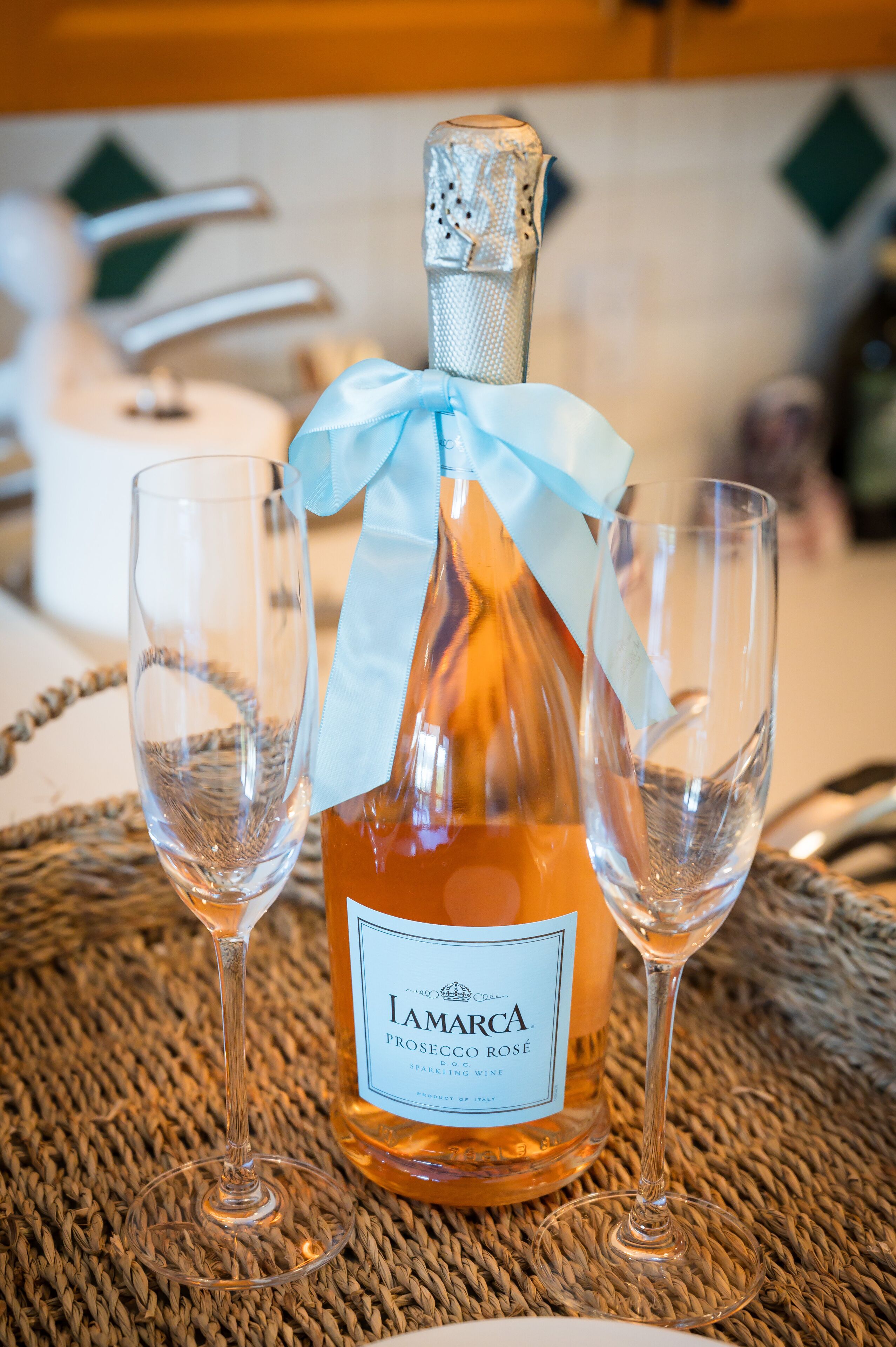 Una bottiglia di Prosecco Rosé con due bicchieri da vino vuoti su un sottopiatti intrecciato.