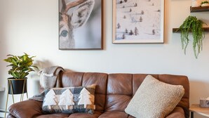 A brown leather sofa with two cushions, a potted plant, and two framed artworks on the wall. - Whistler Village studio, hottub, king bed, free parking (Whistler)