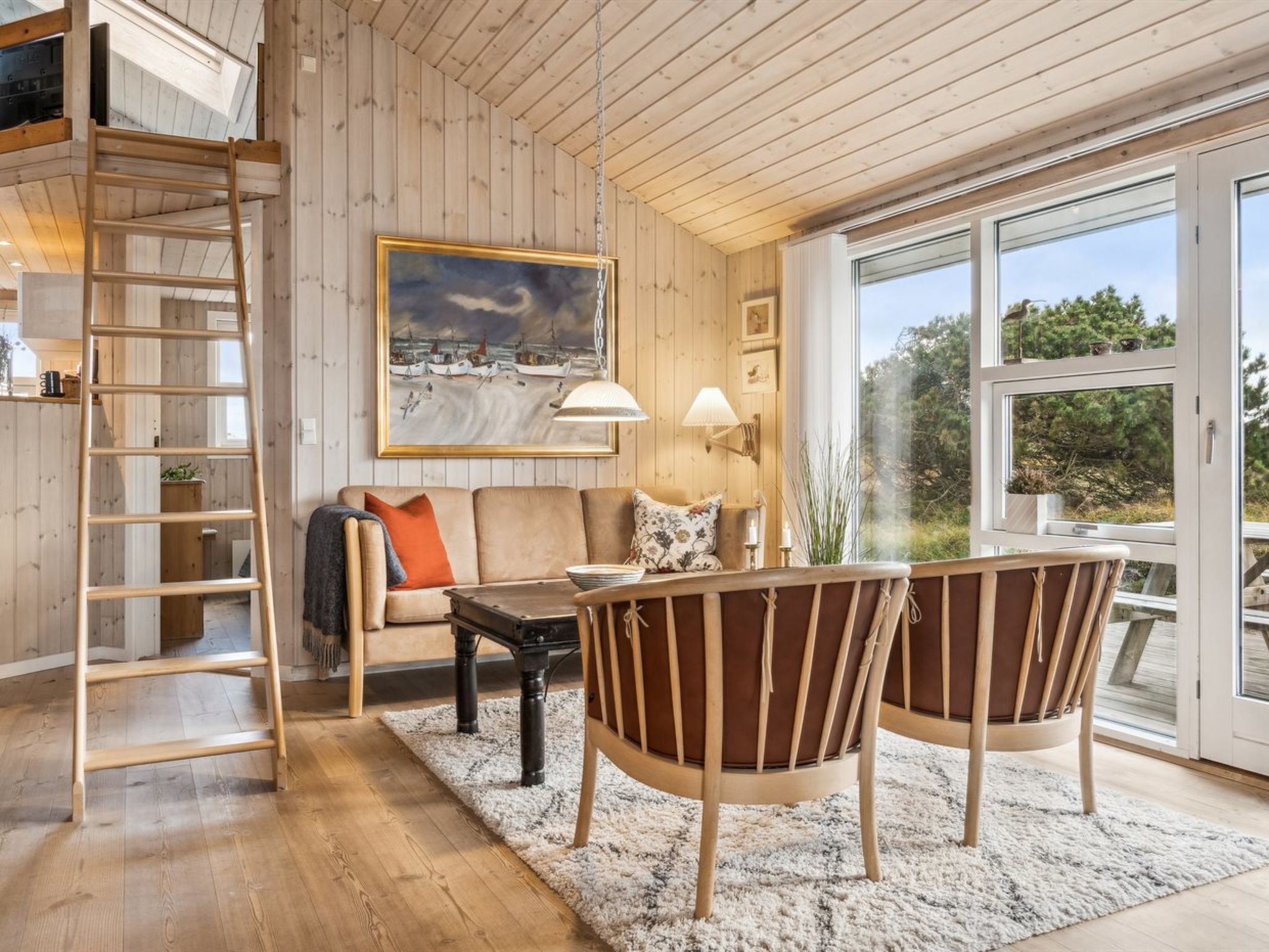 A cozy living room with a wooden floor, a sofa, two chairs, a coffee table, a painting, a ladder, and a large window with a view of greenery.