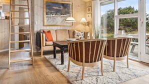 Een gezellige woonkamer met houten vloer, een bank, twee stoelen, een salontafel, een schilderij, een ladder en een groot raam met uitzicht op groen.