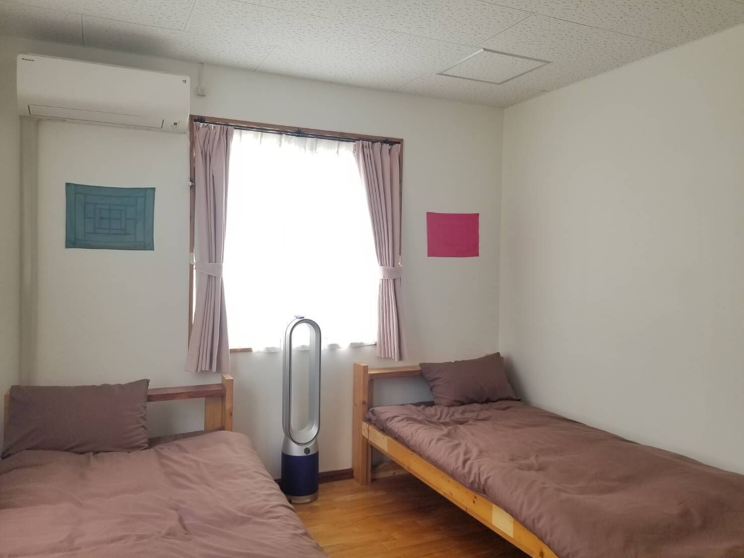 A dormitory room with two single beds, a window with curtains, a radiator, and a small wall decoration.