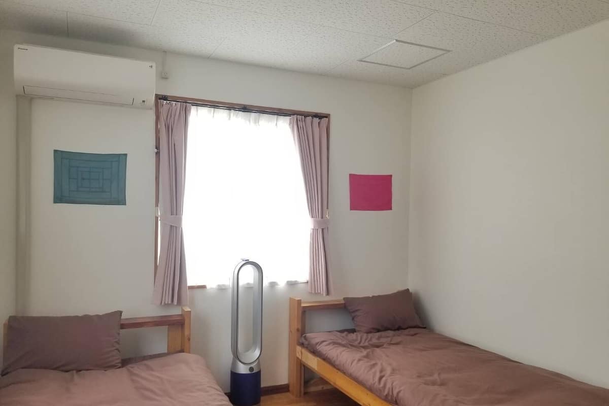 A dormitory room with two single beds, a window with curtains, a radiator, and a small wall decoration.