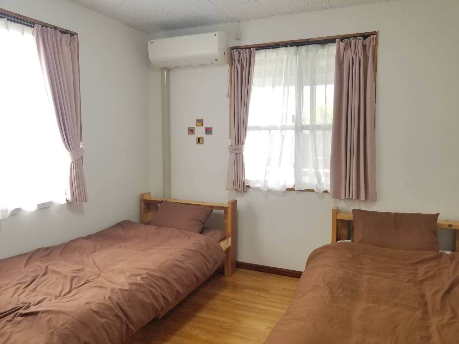 A room with two single beds, a window with curtains, an air conditioning unit, and a small wall decoration.