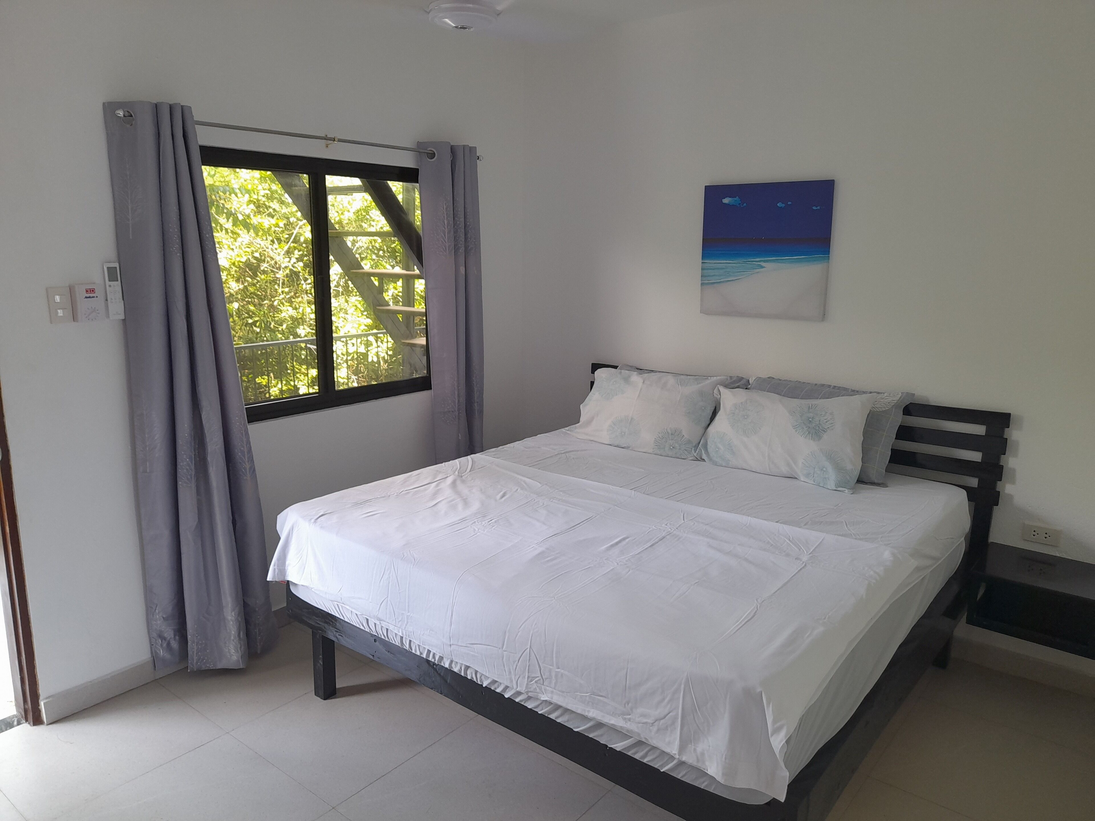 Un dormitorio con cama, cortinas, un cuadro y una ventana con vistas a árboles.