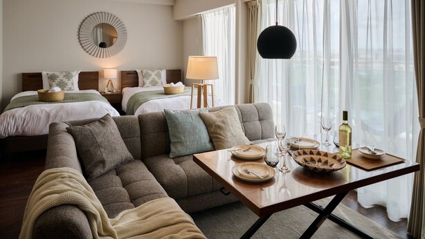 A hotel room with a sofa, two beds, a dining table set with plates, glasses, and a bottle. - Casa YONNA Naha (Naha)