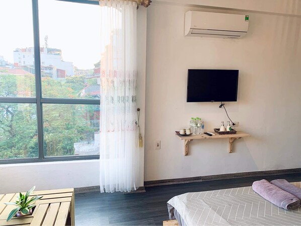 Hotellihuone, jossa on suuri ikkuna, sänky, seinälle kiinnitetty televisio, pieni puinen hylly, jossa on hygieniatarvikkeita, ja ruukussa oleva kasvi.