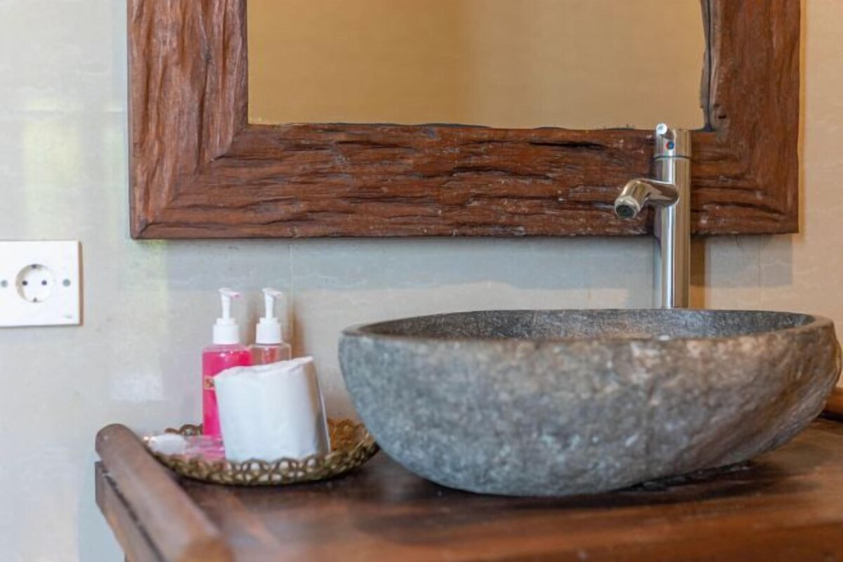 A bathroom with a stone sink, wooden frame mirror, and toiletries on a wooden tray.