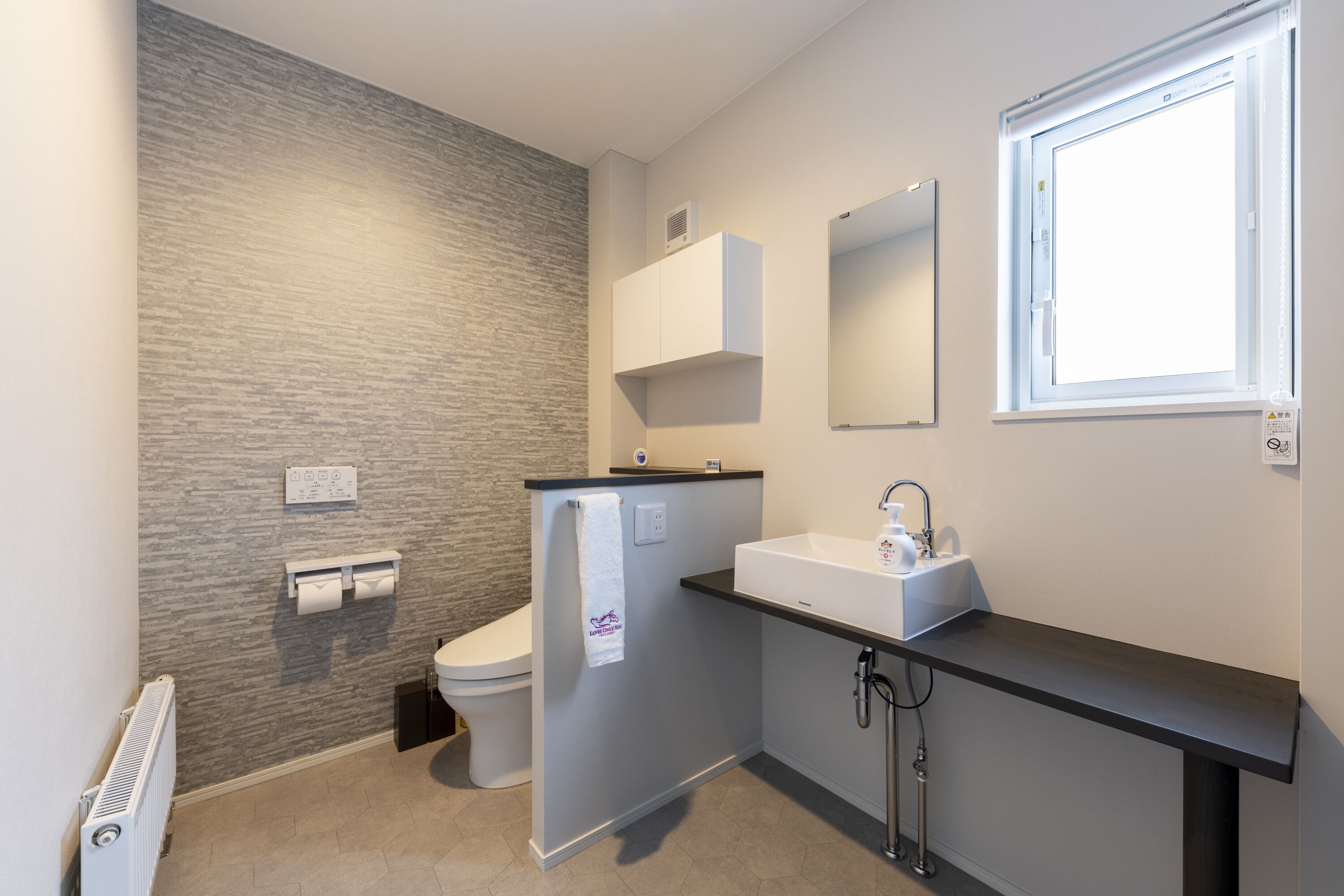 A modern bathroom with a grey textured wall, a white toilet, a black countertop sink, and a window above the sink.
