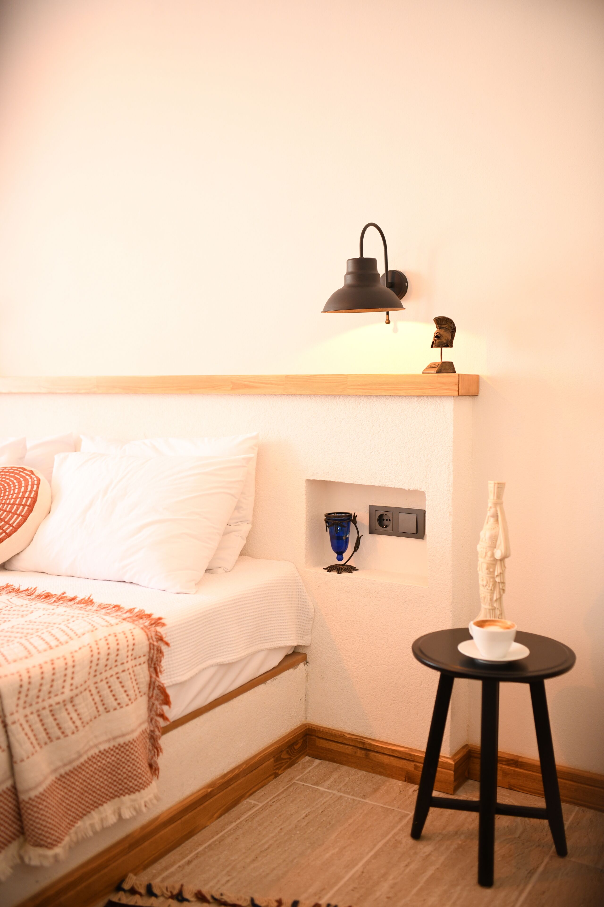 A neatly arranged bedroom with a bed, bedside table, lamp, and a small shelf with decorative items.