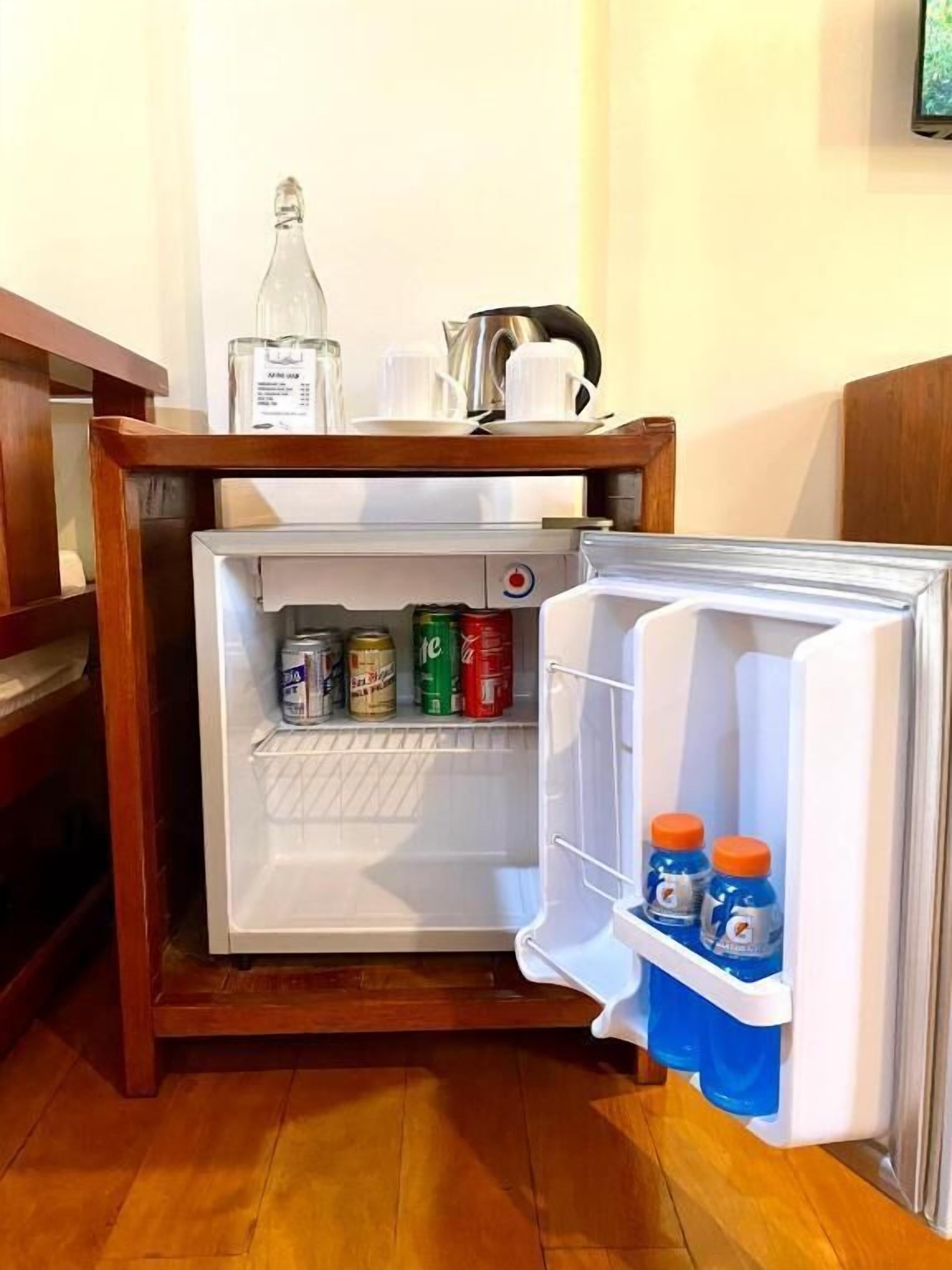 A small refrigerator with a wooden shelf above it, containing a bottle, cups, and a kettle. Inside the fridge, there are cans and bottles of beverages.