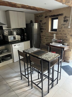 A compact kitchen with a refrigerator, microwave, and coffee maker. There is a dining table with chairs and a small island with a sink. The walls are textured, and there's a window with a view of the outside.