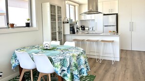 A modern kitchen with white cabinets, a stainless steel range hood, and a built-in oven. There is a dining area with a table covered in a tropical leaf tablecloth and white chairs. - Cosy unit with delightful park and water views. Perfect for a family holiday! (BALLINA)