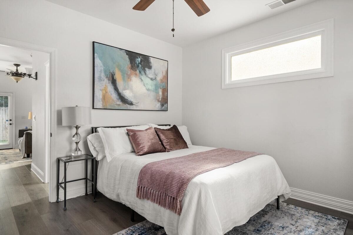 A bedroom with a bed, a nightstand, a lamp, a ceiling fan, a window, and a painting on the wall.