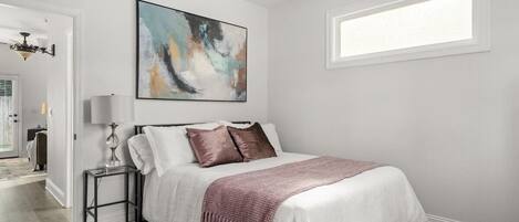 A bedroom with a bed, a nightstand, a lamp, a ceiling fan, a window, and a painting on the wall.