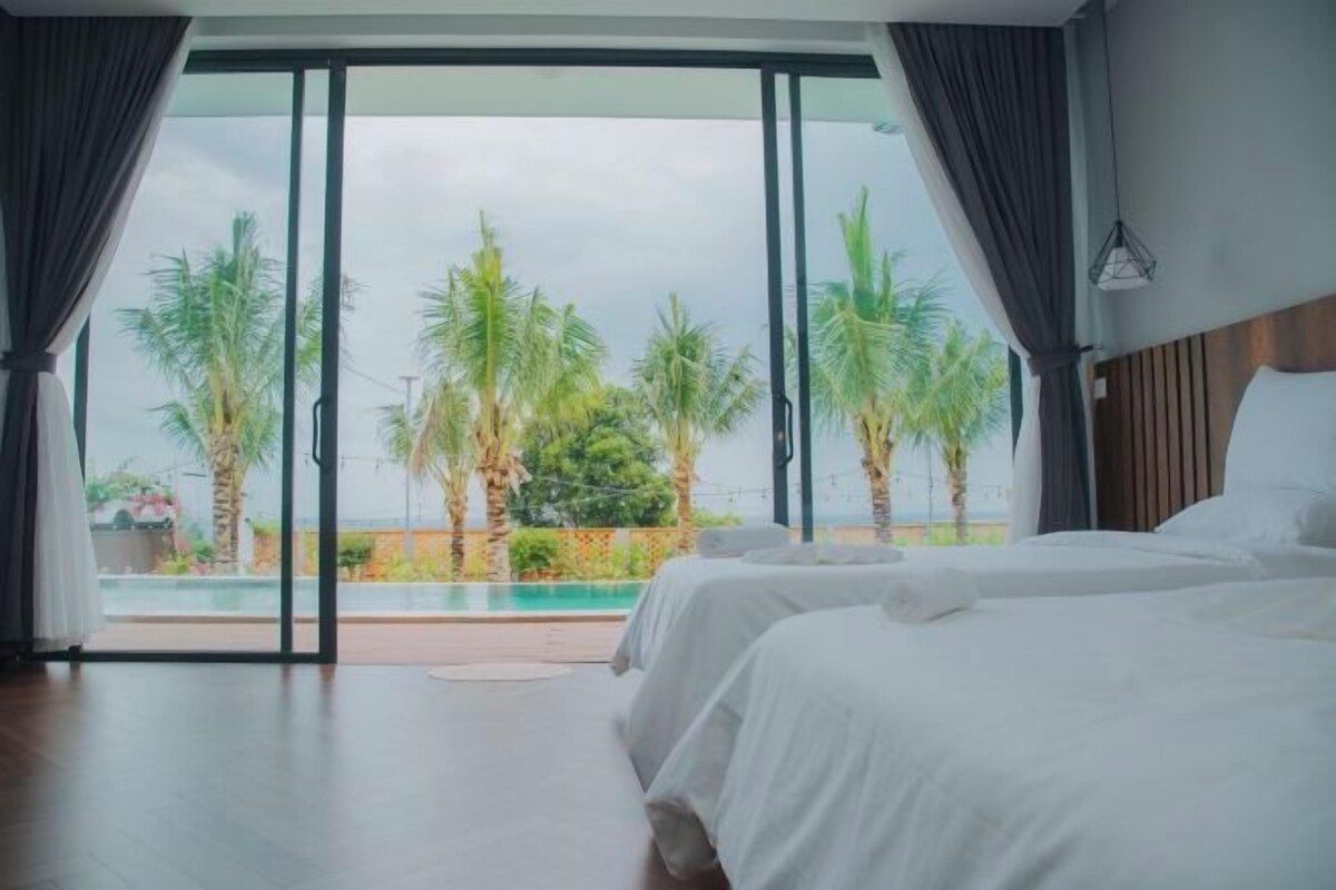 Ein Schlafzimmer mit zwei Betten, einer großen Schiebeglastür, die zu einem Poolbereich führt, und Blick auf Palmen.