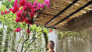 Una dona asseguda al costat d'una banyera en un jardí amb un teulat de bambú i un arbre amb flors roses.