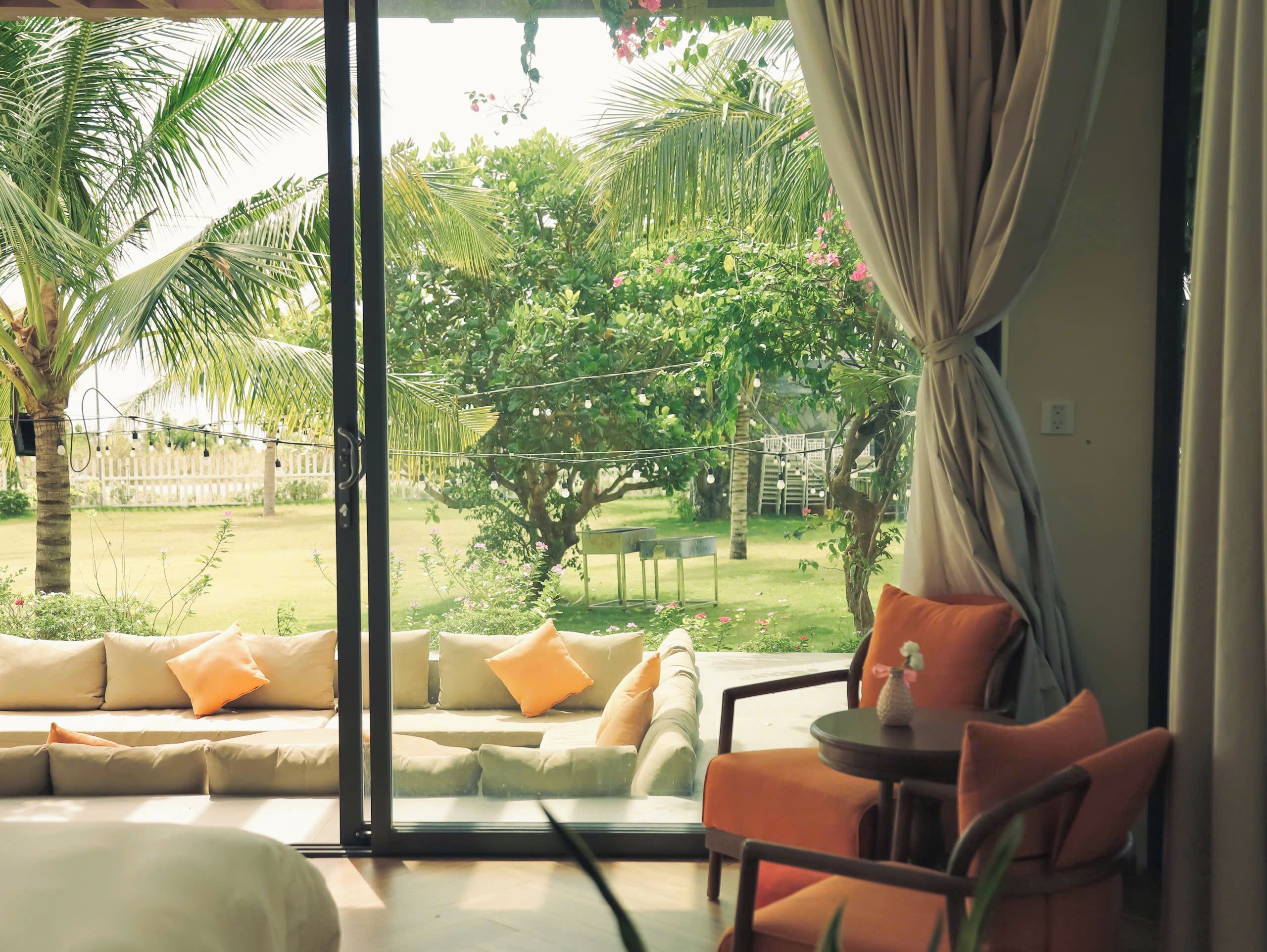 Una habitación con un gran ventanal corredero que da a un jardín. En el interior, hay un sofá beige con cojines naranja, una mesita redonda y una silla.