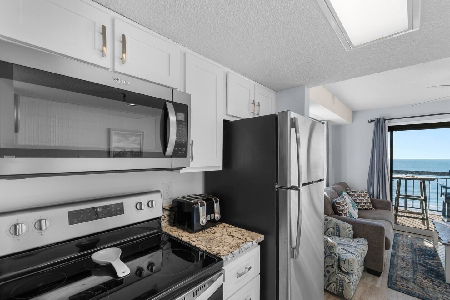 A modern kitchen with a microwave, oven, and refrigerator, a dining area with a table and chairs, and a balcony with a view of the sea.
