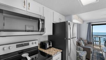 A modern kitchen with a microwave, oven, and refrigerator, a dining area with a table and chairs, and a balcony with a view of the sea.