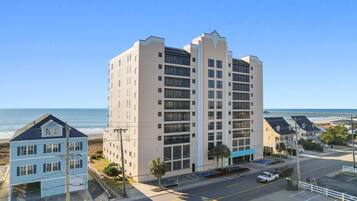 A multi-story hotel with a beach view, parking area, and a pier in the background.