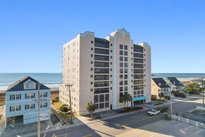 A multi-story hotel with a beach view, parking area, and a pier in the background.